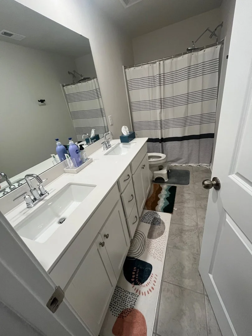 A bathroom with a dual-sink white vanity, large mirror, striped shower curtain, toilet, and multicolored rug on tiled floor.