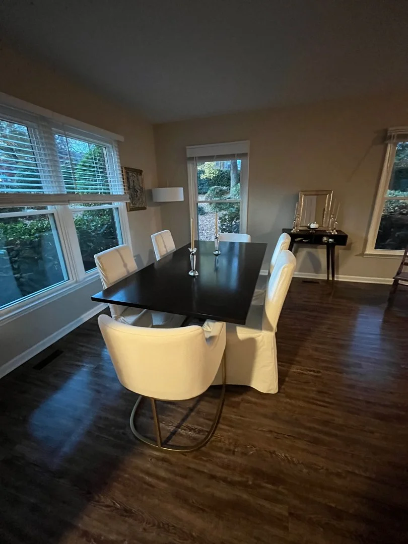 Dining room with dark wooden table, six white chairs, windows with blinds, and a side table with candles and decor