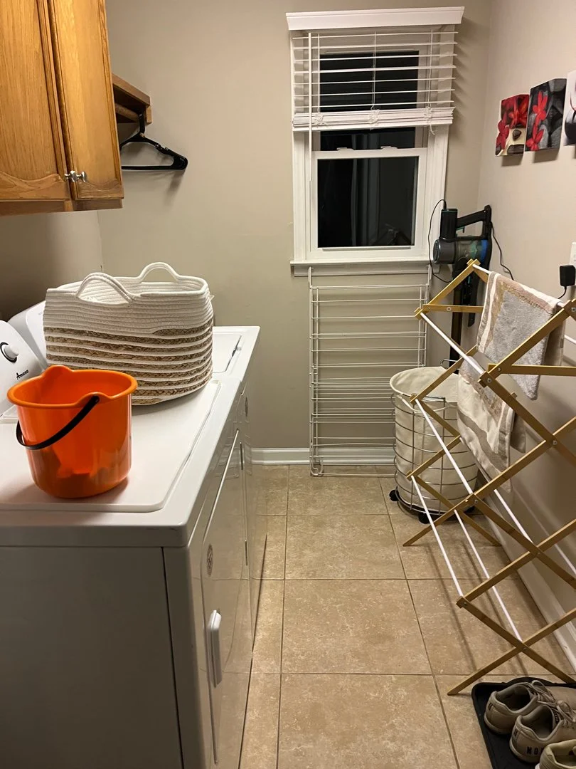 A laundry room with a white washing machine, a stack of woven baskets on top, an orange bucket, a wooden drying rack, a laundry basket, a window with white blinds, a wall-mounted hairdryer, and shoes on the floor.