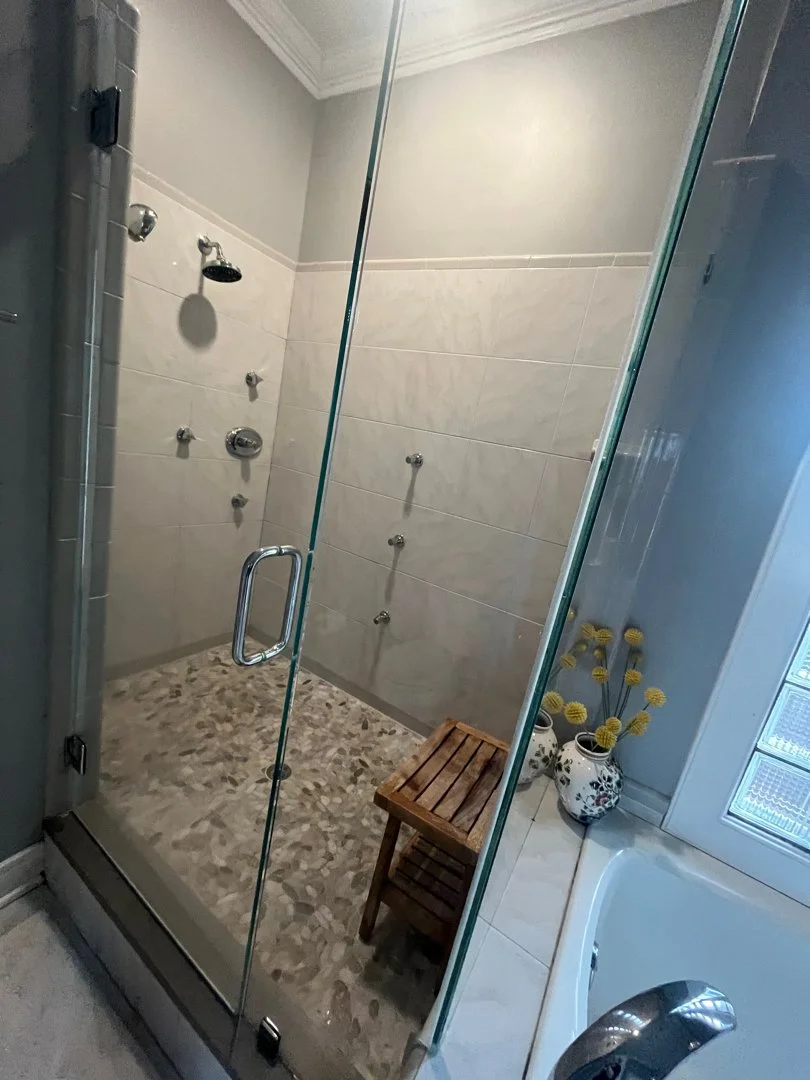 Glass-enclosed shower with beige tiled walls, pebble stone flooring, and a small wooden chair inside. Outside the shower, there are two vases with yellow decorative flowers next to a bathtub with a silver faucet.