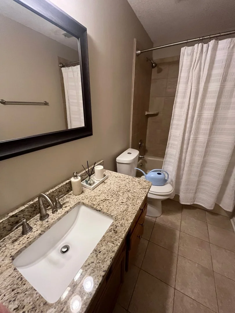 Bathroom with a granite countertop, a white sink, a large mirror, a toilet with a blue watering can on the lid, a shower with a curtain, and beige tile flooring.