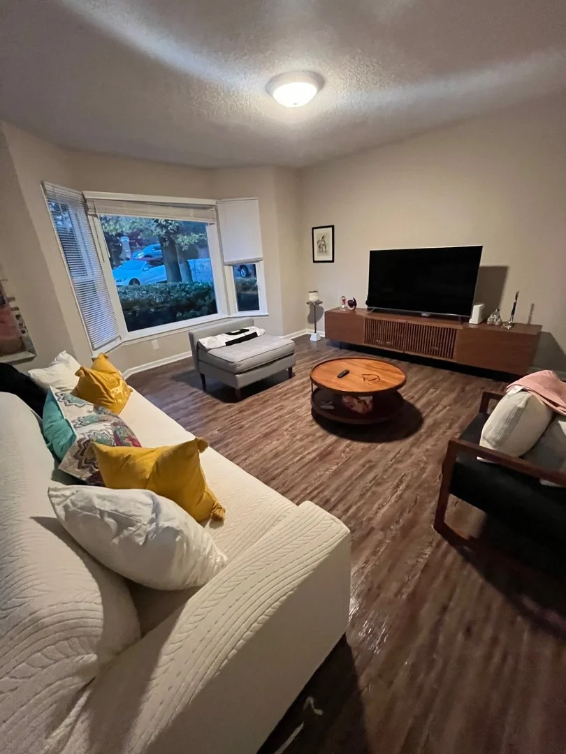 Living room with a cream sofa with colorful pillows, a TV on a wooden stand, a gray armchair, and a round coffee table on wood flooring. There are large windows with blinds, a lamp, and framed artwork on the wall.