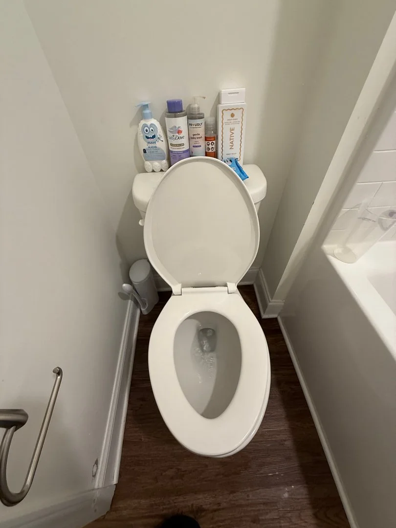 Toilet with the lid open in a bathroom, with toiletries placed on top of the toilet tank, including bottles of soap, lotion, and other personal care products, next to a bathtub and a support grab bar.