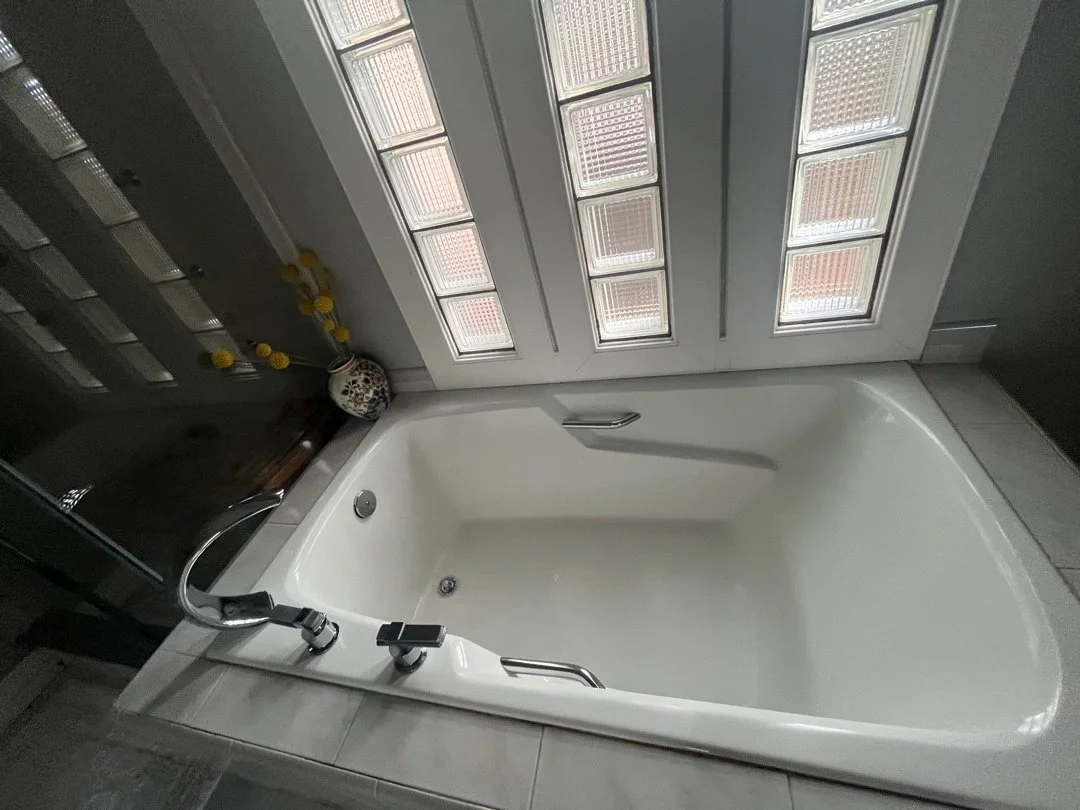 Empty white bathtub with metal handles, located in front of a window with blinds, with a decorative vase and yellow flowers nearby.