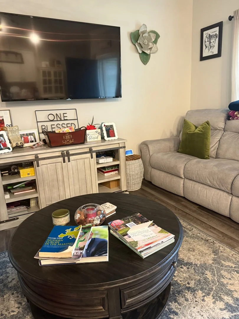 Living room with a large flat-screen TV mounted on the wall, a gray couch with a green pillow, a round black coffee table with magazines and a glass jar with candles and decorations, a white distressed sideboard with picture frames, plants, and decorative signs, and artwork on the wall.