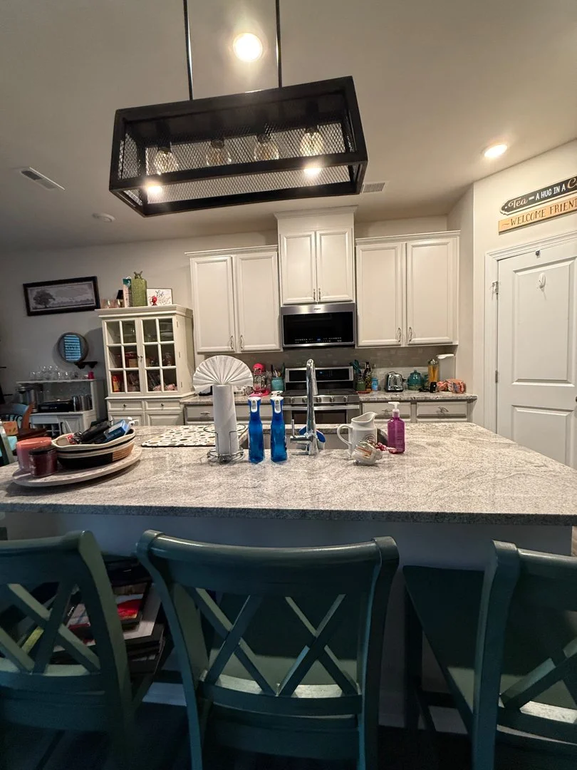 A kitchen with a large granite island counter, three barstools, white cabinets, and various kitchen appliances and items. Overhead lighting fixture is visible.