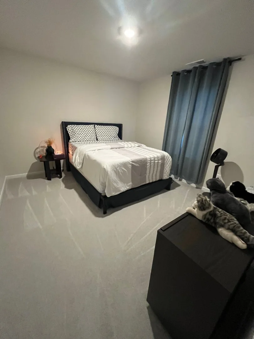 A minimalist bedroom with a bed with white bedding, black headboard, and pillows; a small black side table with a black vase and dried flowers; dark gray curtains on a window; a black dresser with plush stuffed animals on top; and gray walls with a c