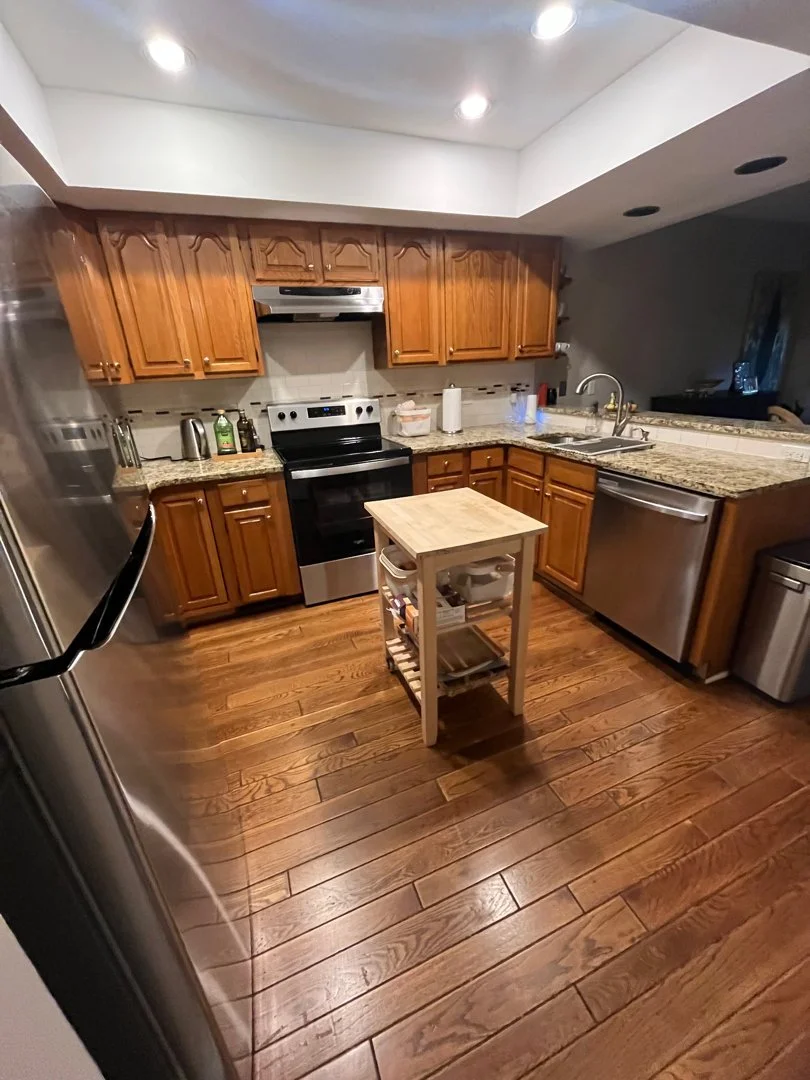 Kitchen with wooden cabinets, stainless steel appliances, and a small mobile kitchen island on hardwood flooring.