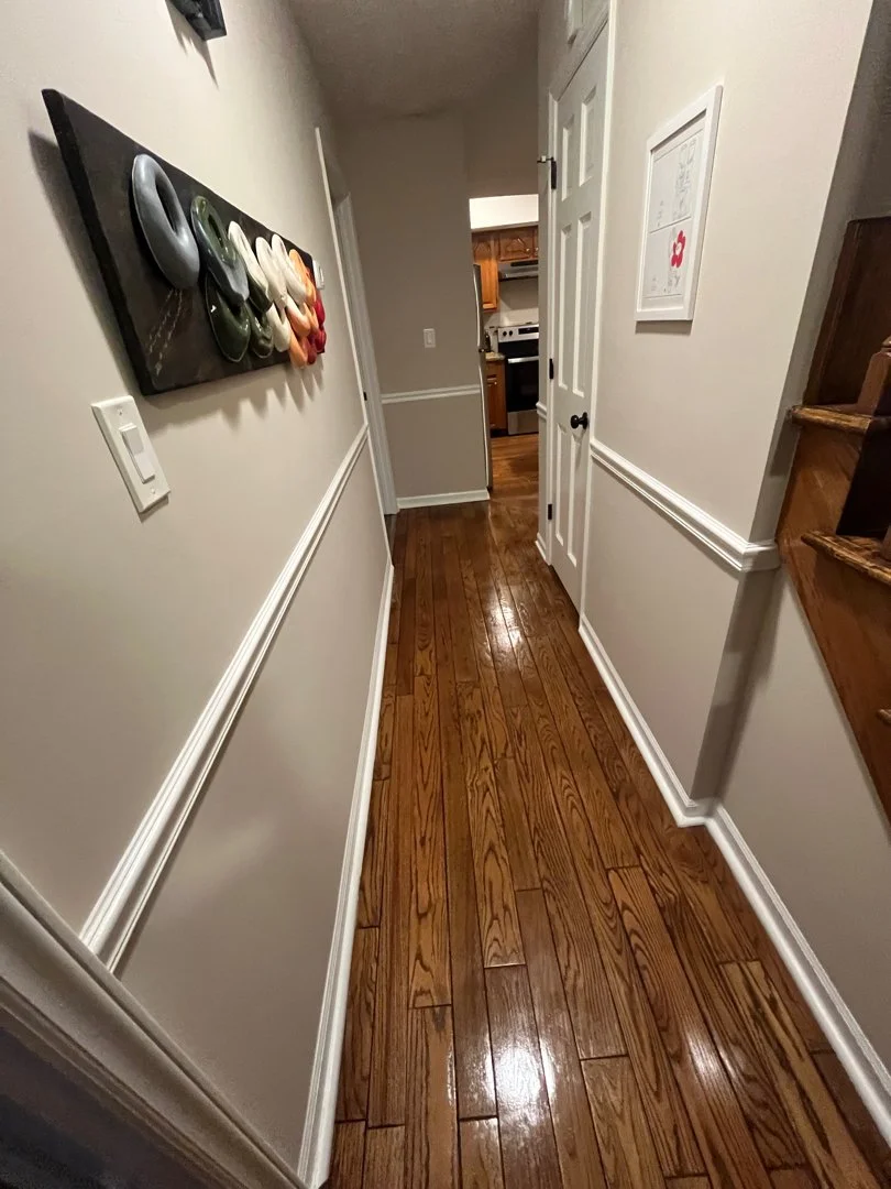 Photo of a hallway with wooden floors, white walls, and white trim. There are two doors on the right side—one open leading to a kitchen with brown cabinets and a white stove. The left side has a wall hanging with decorated stones and a switch. There 