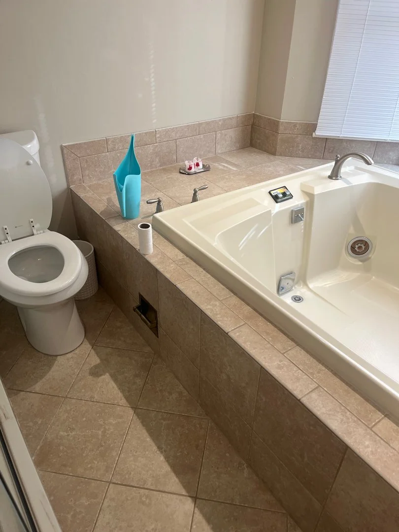 Bathroom with tiled floor and includes a toilet, a large bathtub, and various cleaning accessories like toilet paper, a blue spray bottle, and soap dishes.