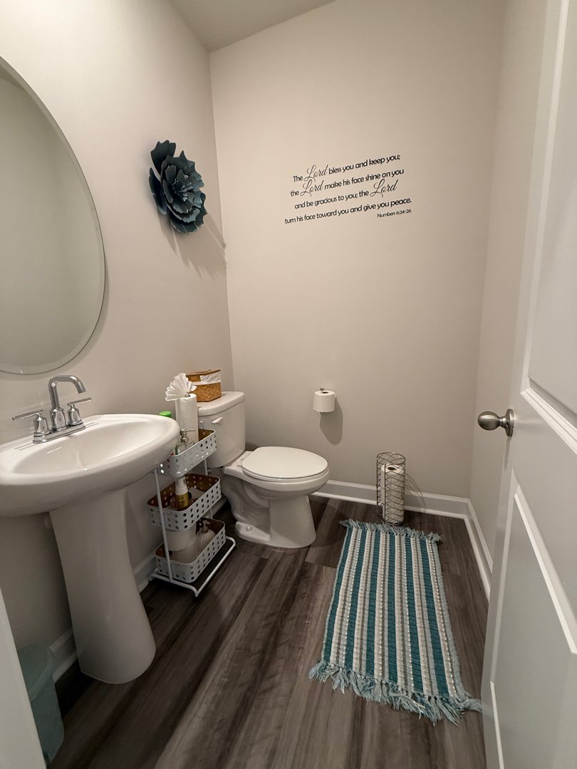 A small, clean bathroom with a white pedestal sink, a round mirror, a toilet with a tissue box on top, a small white shelf holding toiletries, a teal and beige striped rug, a white wall decal with a scripture, a blue flower wall decoration, a white d