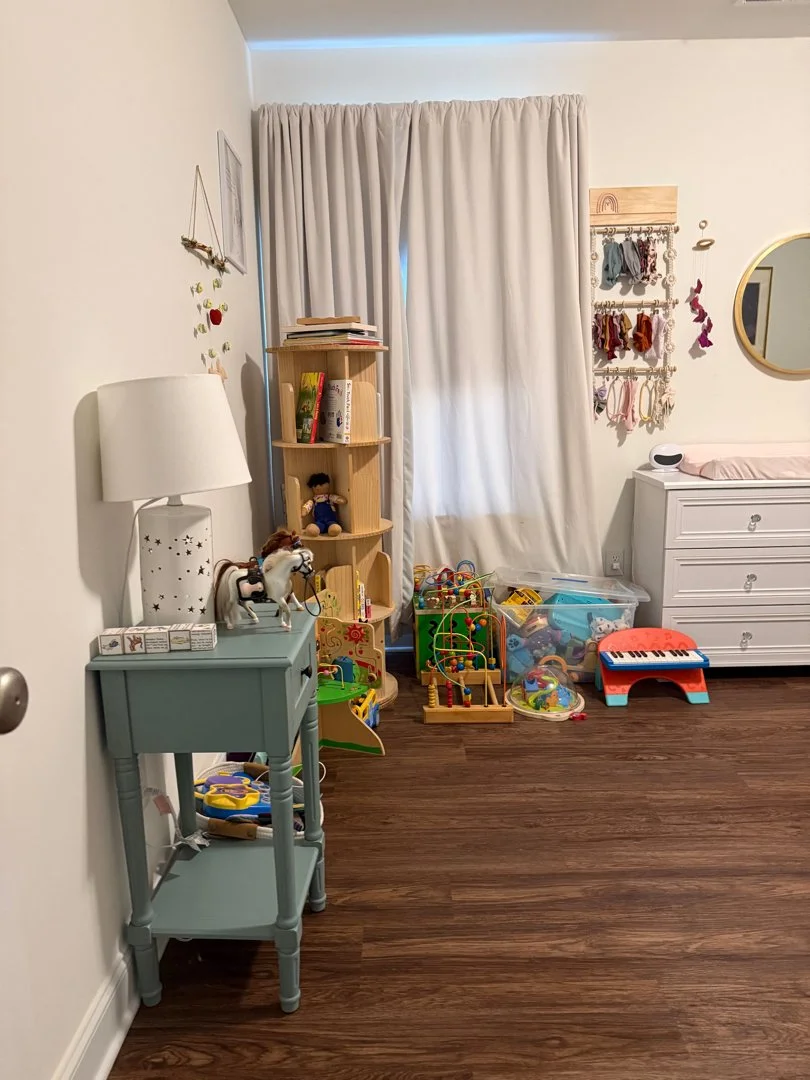 Child's playroom with a blue side table holding a lamp and toy horse, a wooden bookshelf with books and a doll, a wall with hanging decorative items, a large window with beige curtains, and colorful toys on the floor including a plastic keyboard and 