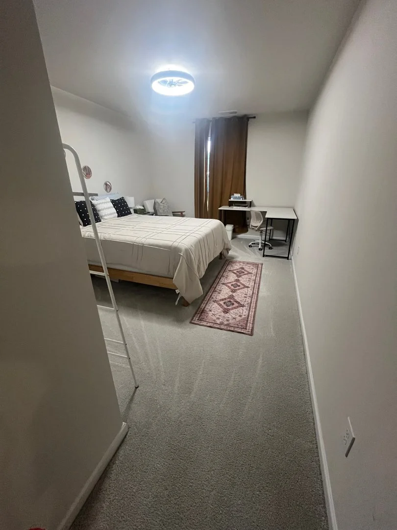 A minimalist bedroom with a bed, side table, chair, desk, and window covered by brown curtains.