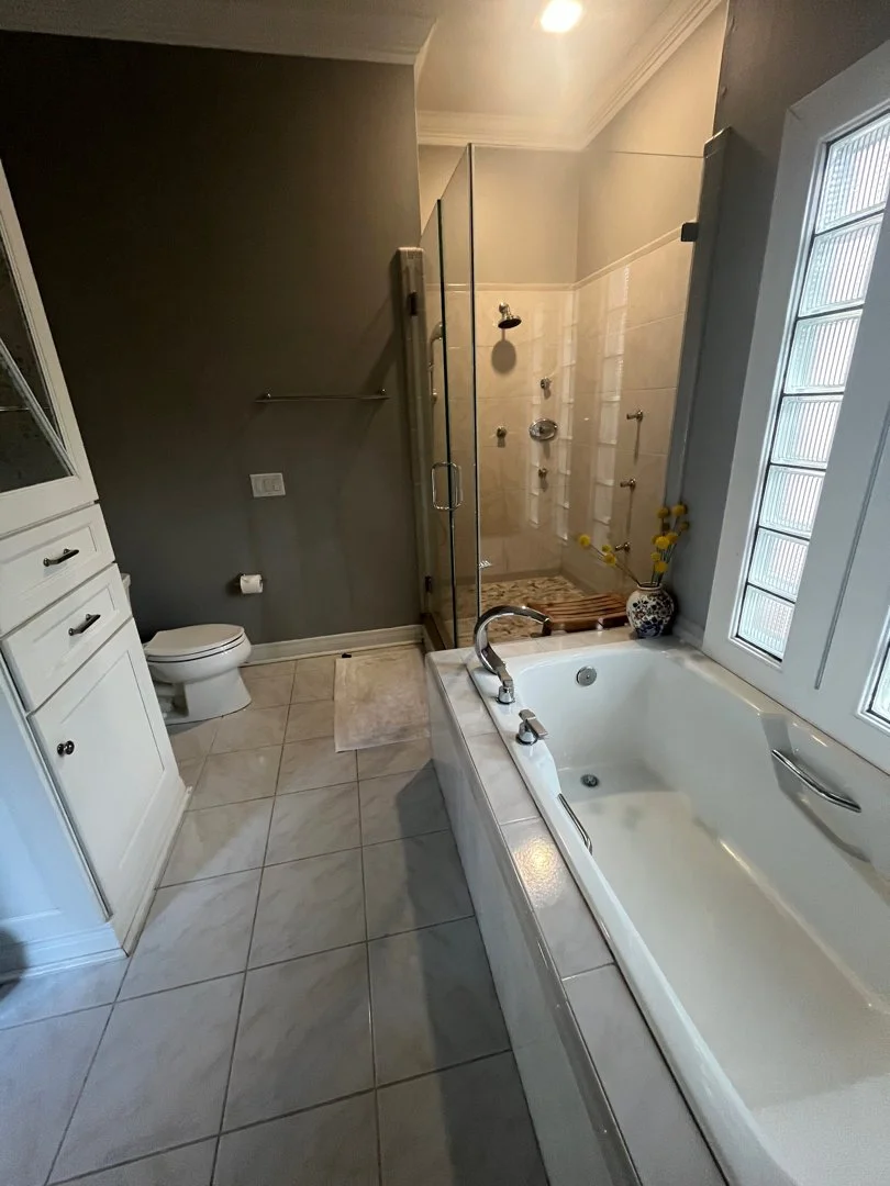 A bathroom with a toilet, a glass shower enclosure, a bathtub with a window nearby, and white cabinets, with beige tiles on the floor and a dark gray wall.