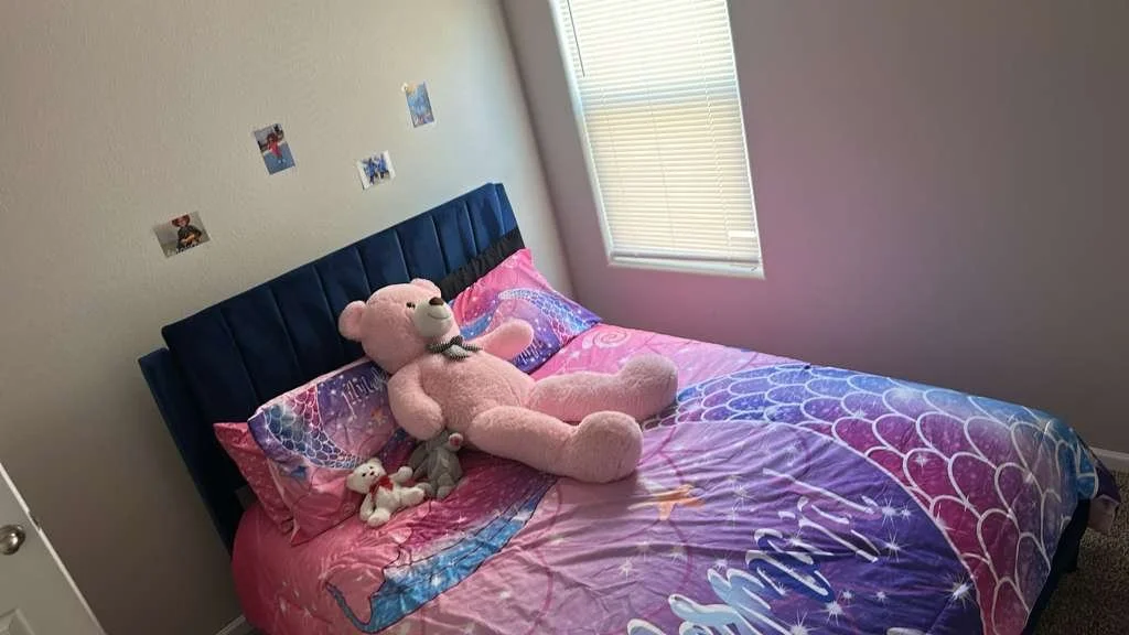 A cozy child's bedroom featuring a bed with pink and purple bedsheets decorated with a mermaid and scales pattern. On the bed, there's a large pink teddy bear, a smaller white plush toy, and a gray stuffed animal. The wall above the bed has small pho