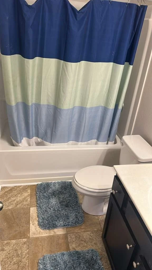 Bathroom with a shower curtain, toilet, dark vanity with a light countertop, and two blue fuzzy rugs on a tan tiled floor.