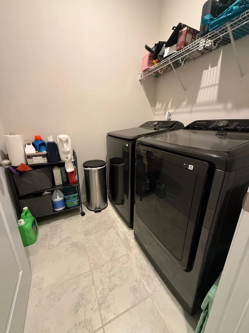 A laundry room with a black washing machine and dryer, a wire shelf above holding various items, and a small black shelving unit with laundry supplies and paper towels.