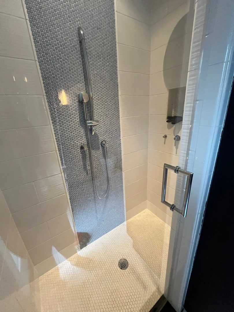 A modern shower area with gray hexagon tile floor, tiled walls, a handheld showerhead, and a glass door with a metal handle.