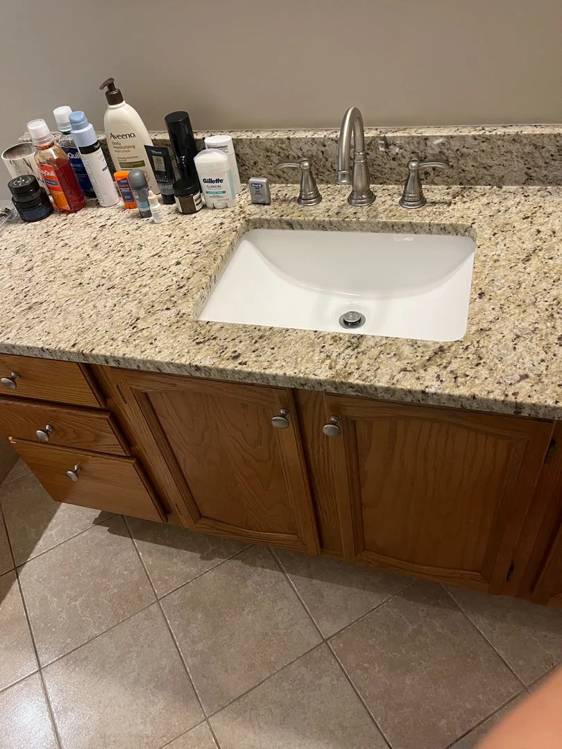 Bathroom countertop with various personal care products, including lotions, sprays, and hygiene items. There is a white sink with a silver faucet and wooden cabinet drawers below. The countertop is speckled granite and the floor has beige ceramic til