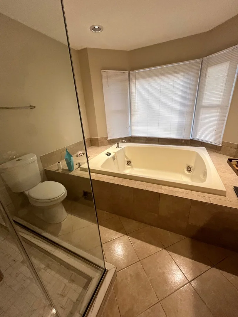 Bathroom with a large corner bathtub, a glass-enclosed shower, and a toilet. The room has beige tiles and a window with blinds.