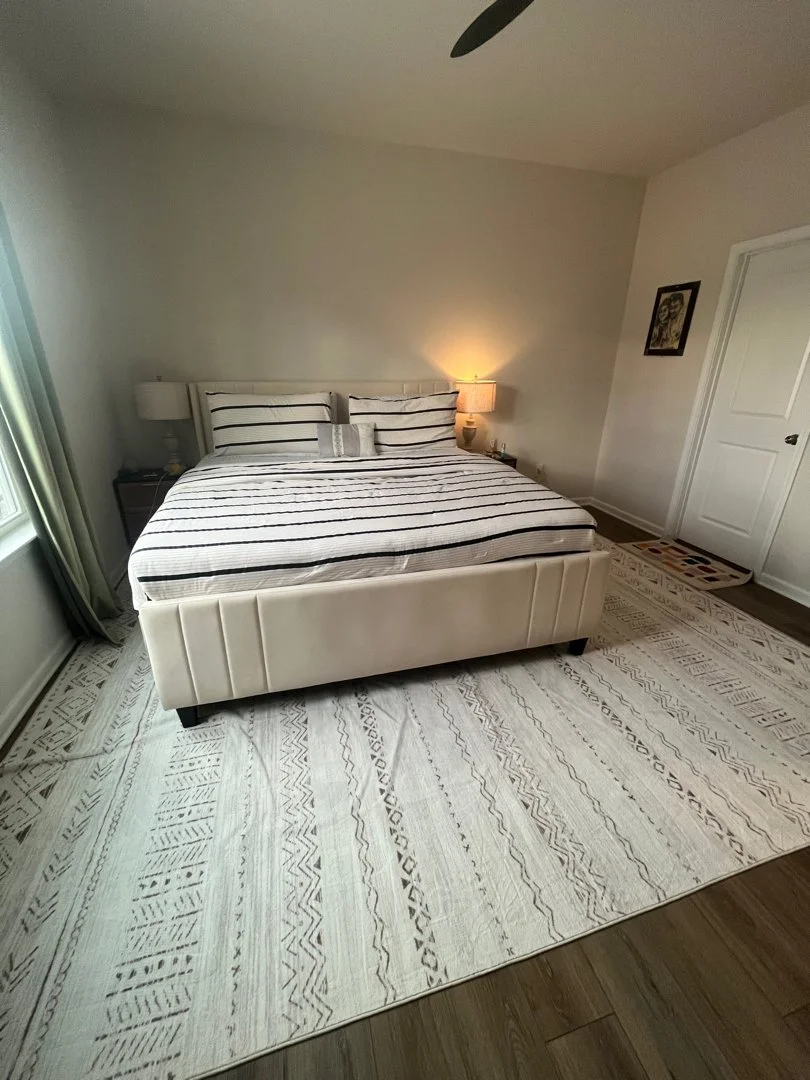 A bedroom with a king-sized bed, striped bedding, and matching pillows. There are nightstands with lamps on each side of the bed, a window with curtains on the left, a ceiling fan above, and a patterned area rug on the wooden floor. A small framed pi