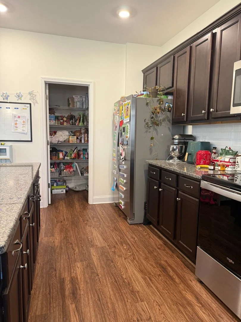 Kitchen with dark wood cabinets, granite countertops, a stainless steel refrigerator with photos and magnets, a microwave, stand mixer, and stove. An open pantry on the back wall stocked with food. Wooden-style flooring.