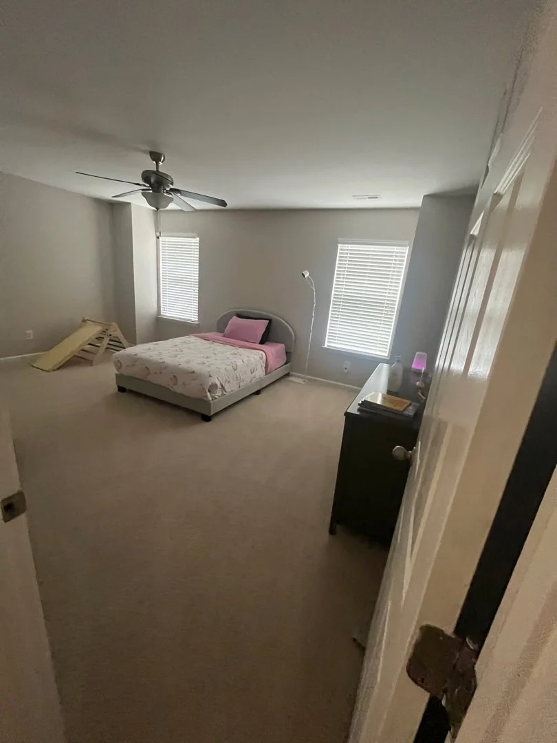 A bedroom with light-colored walls and carpet. It contains a bed with a floral comforter and pink bedding, a black dresser with books and a diffuser, two windows with blinds, a ceiling fan, a floor lamp, and a damaged wooden chair on the floor.