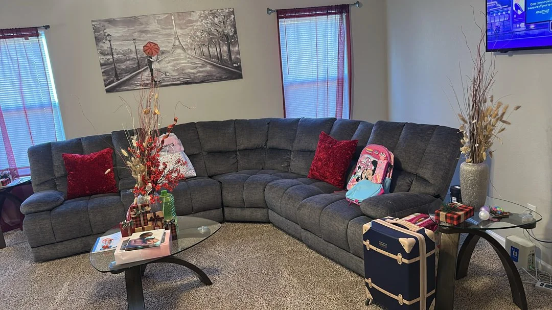Living room with a dark gray sectional sofa, red pillows, and holiday decorations on tables, including a large vase with dried flowers, a suitcase, a backpack, and a flat-screen TV on the wall.