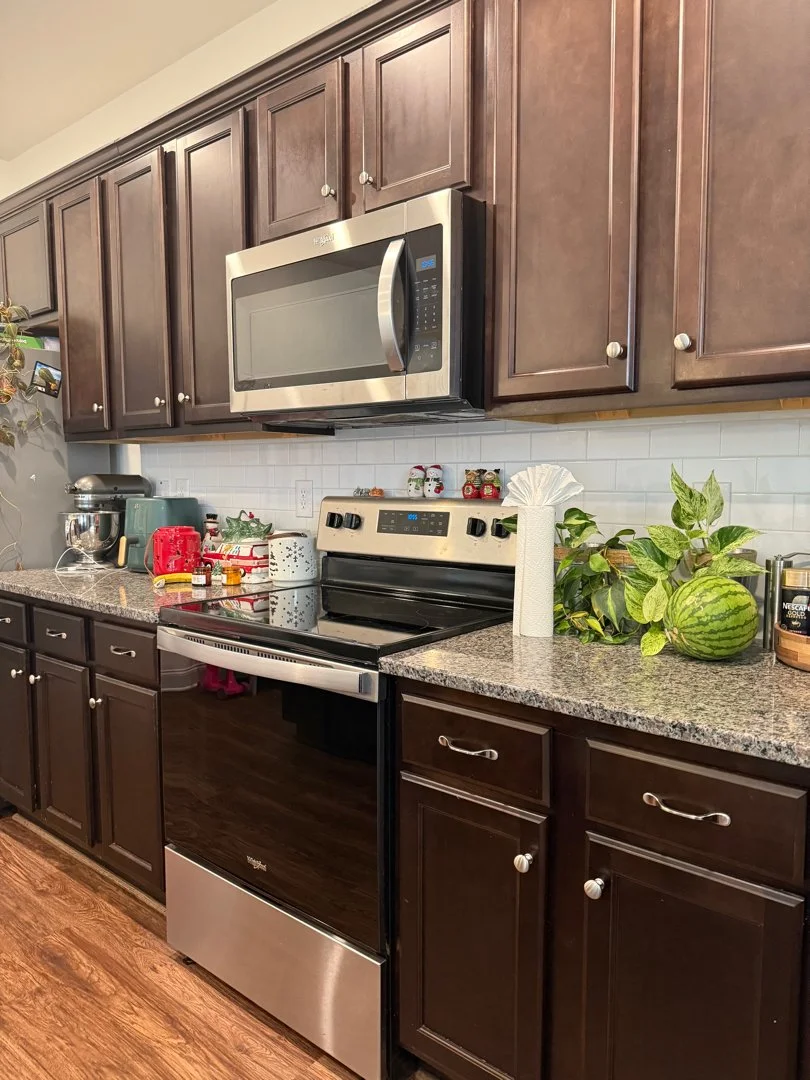 A kitchen with dark wood cabinets, a stainless steel microwave above a black and stainless steel oven, and a granite countertop with various kitchen items and decorative plants.