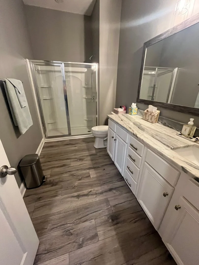 A modern bathroom with gray walls, a white vanity with a marble countertop, a large mirror, a glass-enclosed shower, a toilet, a small trash can, and a towel hanging on the wall.