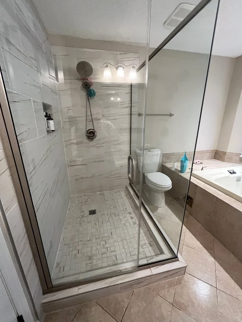 Bathroom with a glass-enclosed shower, a toilet, and a bathtub. The shower has marble tiles, a showerhead, and toiletries in a niche. The toilet is next to the shower, and the bathtub is to the right with a blue container and toiletries on the edge. 