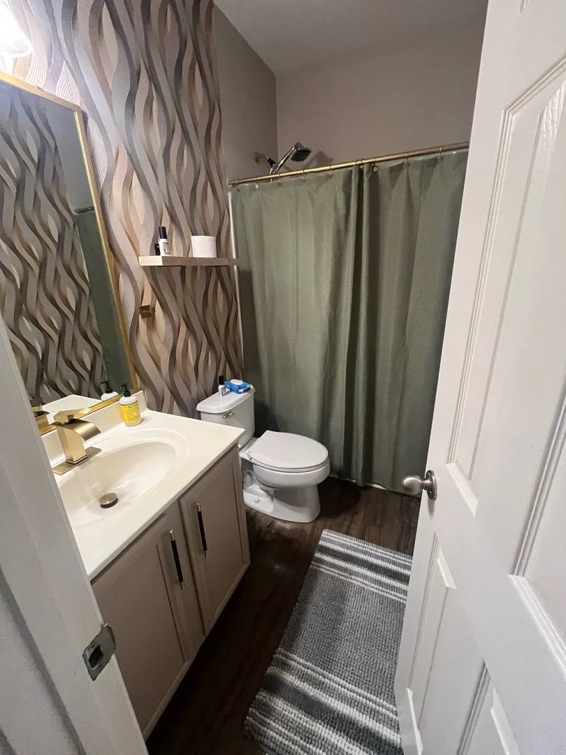 Small bathroom with a beige vanity and sink, gold faucet, wall-mounted mirror, patterned wallpaper, shelf with toiletries, toilet next to shower with green curtain, gray and white striped rug on dark wood floor.
