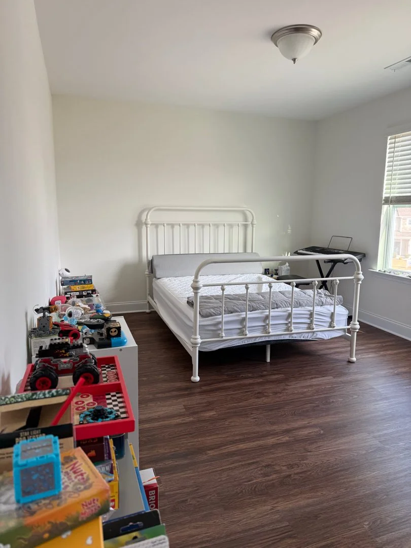 Empty bedroom with a white metal bed frame, a crib with stuffed animals and toys on a nearby shelf, a window with blinds, and a ceiling light fixture.