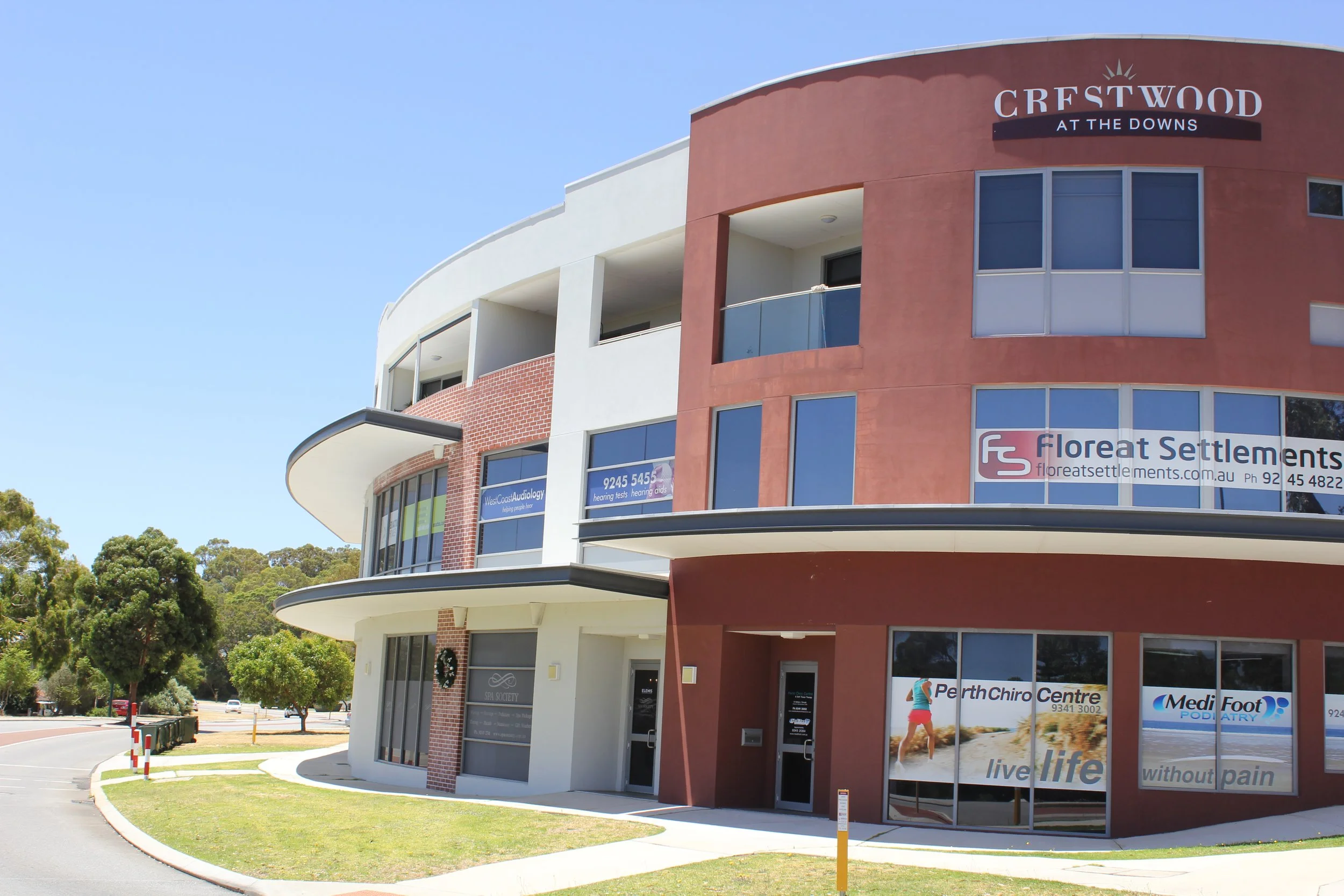 A multi-story commercial building with a curved design, featuring a red brick section and white and painted orange facades, with multiple glass windows and signs for businesses, including Floreat Settlements and PerthChiro Centre.
