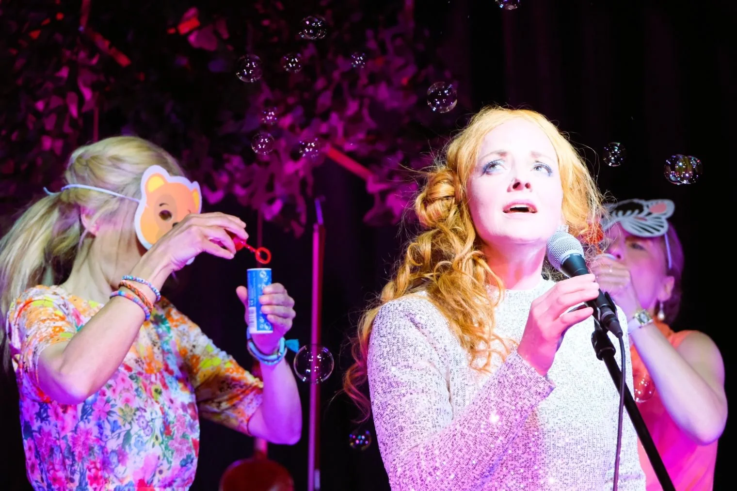 Drei Frauen auf einer Bühne mit Seifenblasen, eine singt mit einem Mikrofon, die anderen zwei tragen Tiermasken und blasen Seifenblasen.