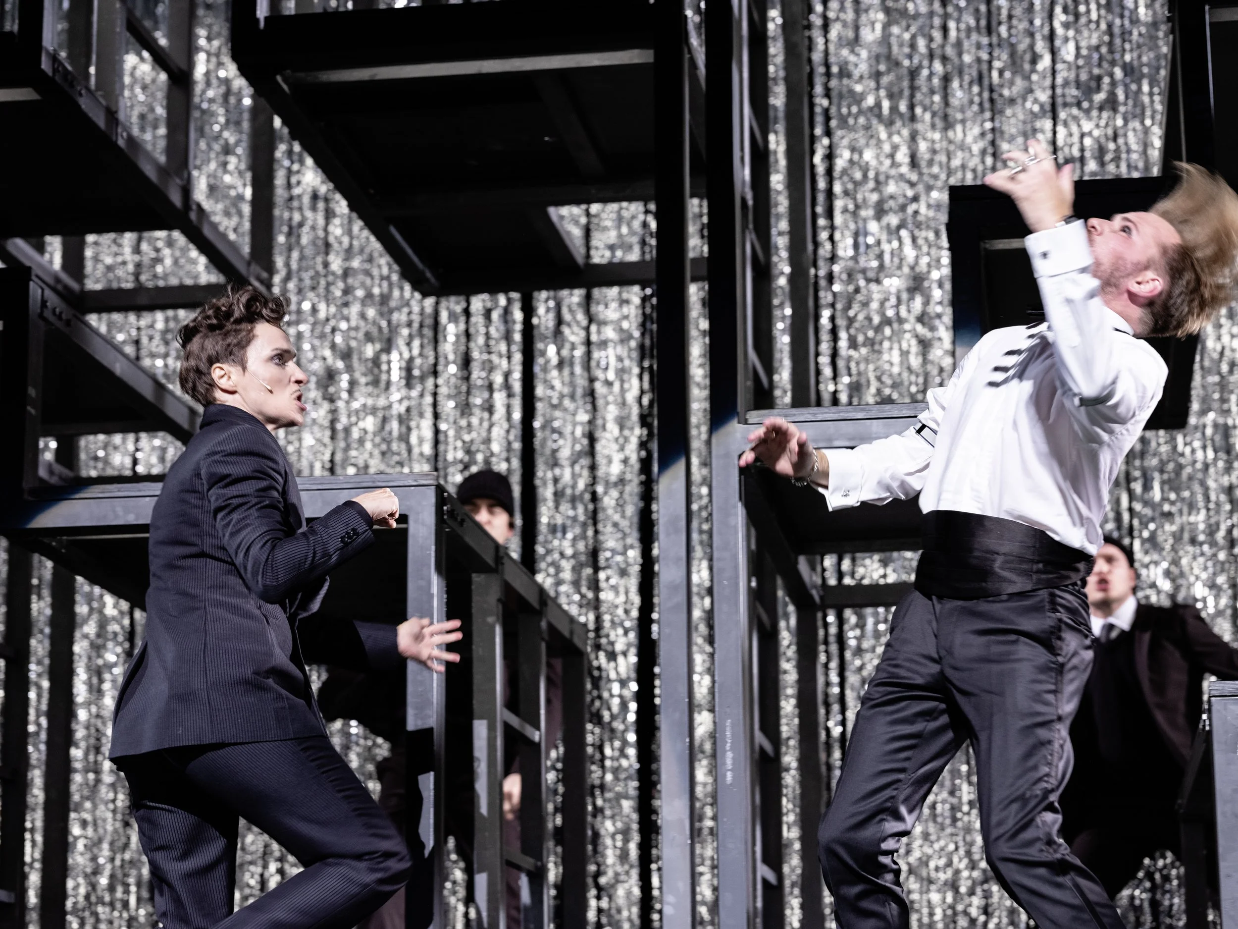 Zwei Männer in formeller Kleidung in einem Bühnenkonflikt, mit einem silbernen, reflektierenden Hintergrund.
