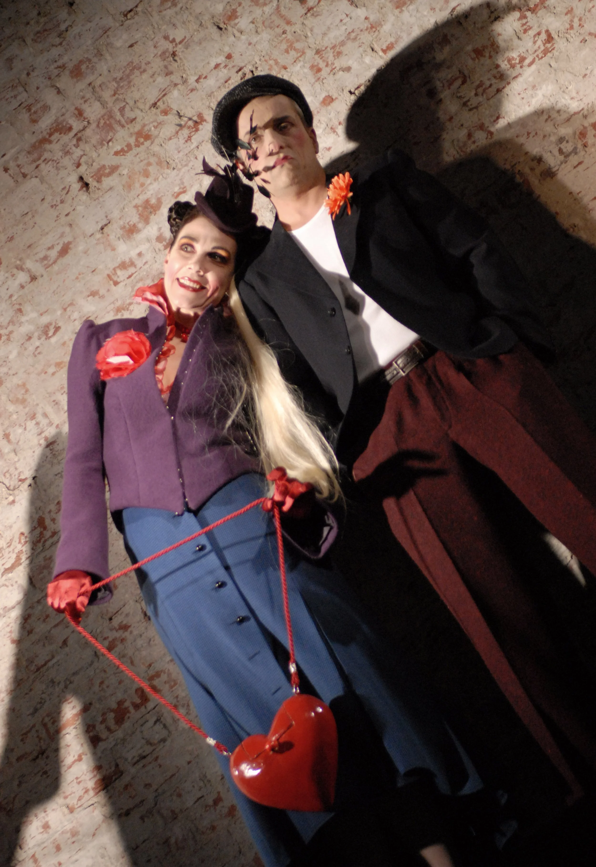 Zwei Menschen in Vintage-Kostümen posieren vor einer roten Backsteinwand, die Frau trägt einen lilafarbenen Mantel mit rotem Herz-Anhänger und die man trägt einen schwarzen Hut, schwarze Jacke und braune Hosen.