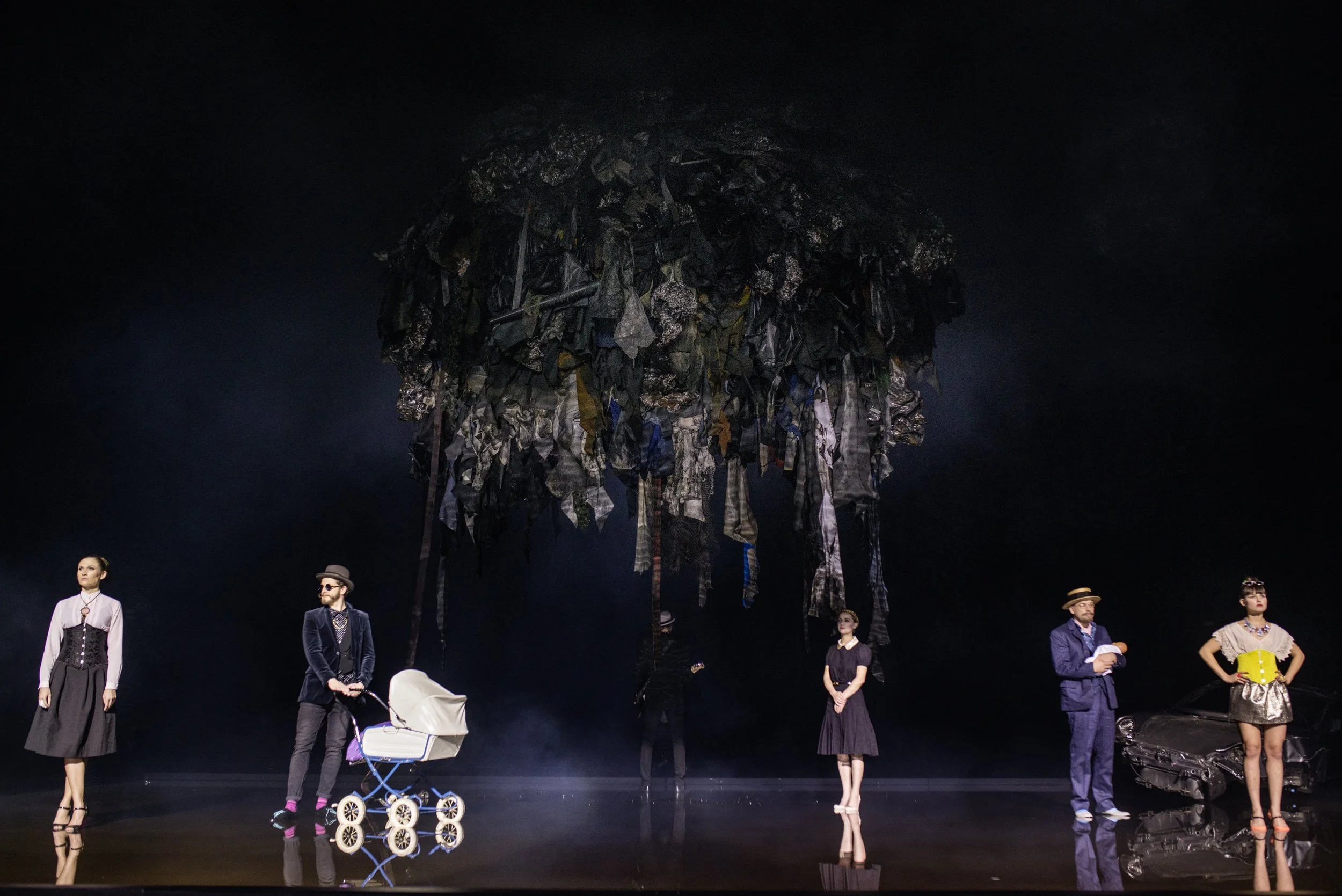 Szene aus einem Theaterstück mit sechs Schauspielern auf einer Bühne, im Hintergrund ein großer Haufen aus zerfetzten Kleidung und Müll, symbolisch für Umweltverschmutzung.
