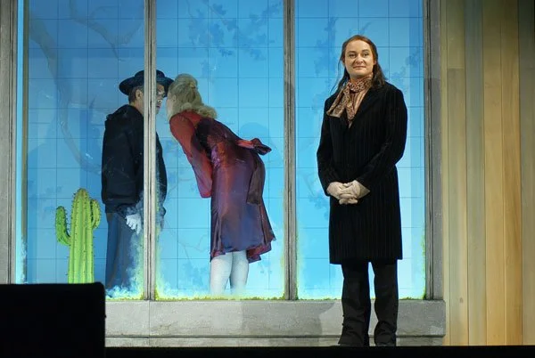 Eine Theaterszene mit drei Figuren, zwei im Fenster und eine außerhalb. Die beiden Figuren im Fenster scheinen sich zu küssen, während die dritte Frau außerhalb des Fensters zuschaut.