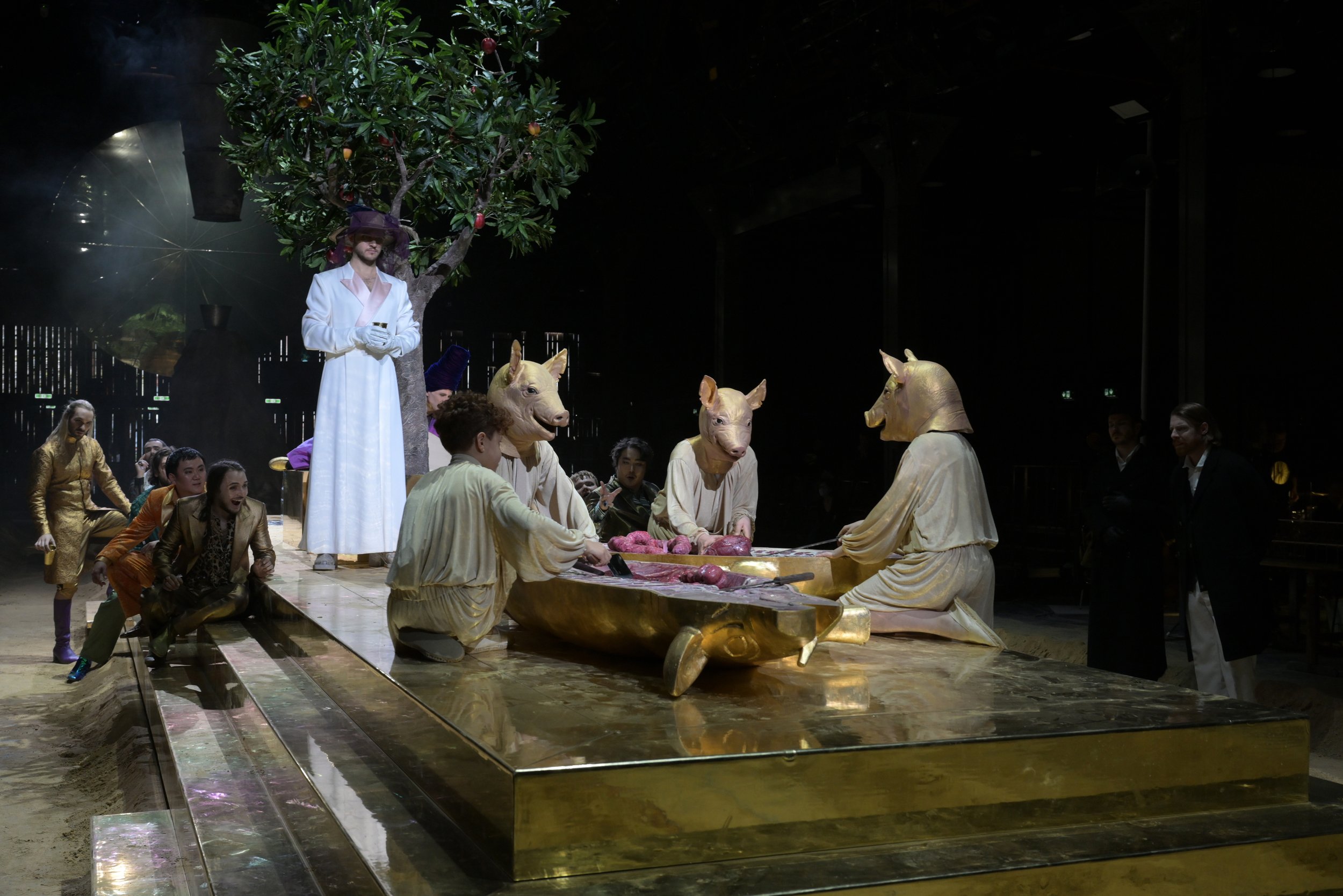 Theaterperformance mit Maskenträgern in Schweine- und Pferdekostümen, auf einer goldenen Plattform, im Hintergrund ein Apfelbaum, Zuschauer und Regiepersonal beobachten die Szene.