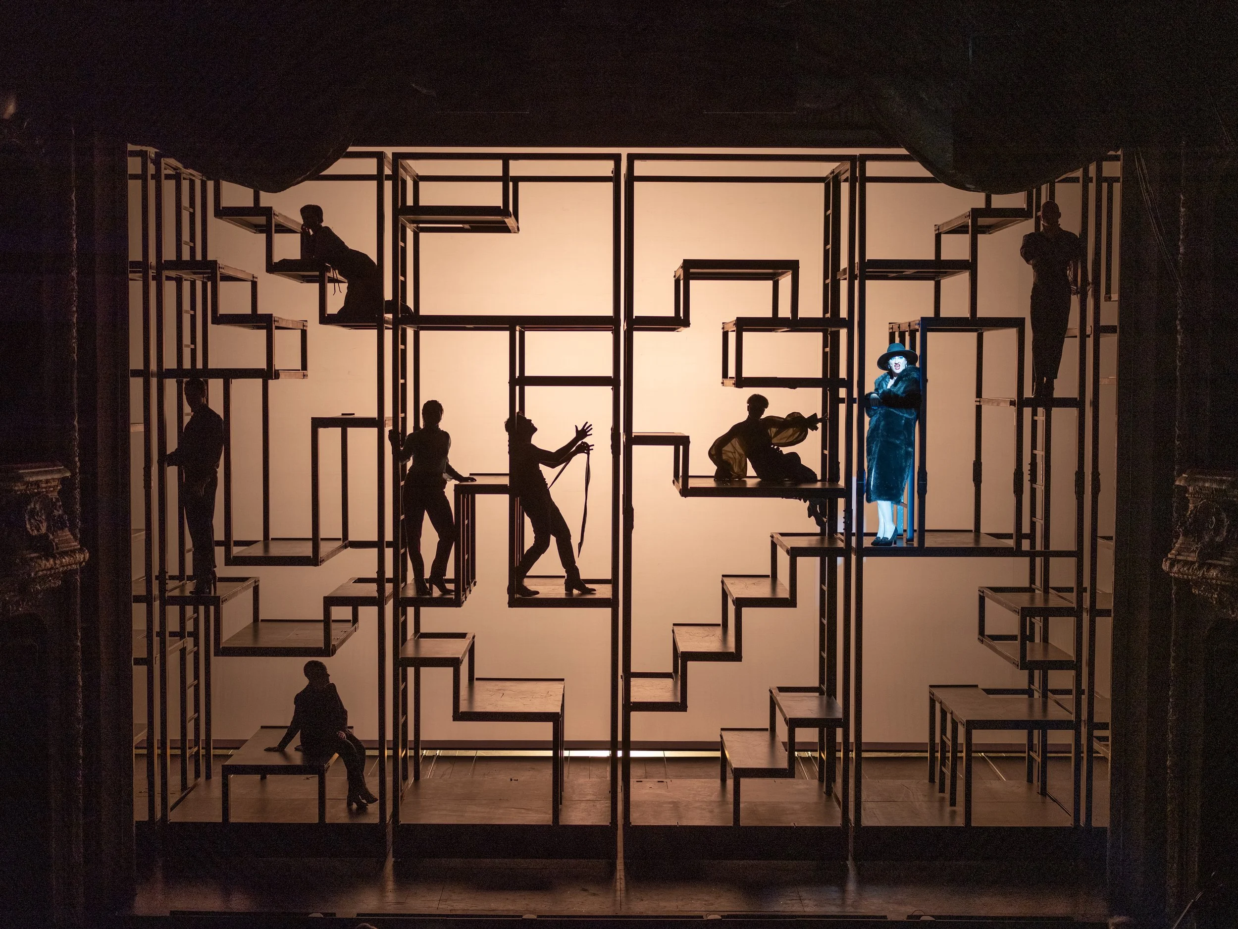 The image shows a theatrical stage set with silhouettes of mannequins and a woman in a dark dress and hat, lit from behind, surrounded by geometric metal frames.