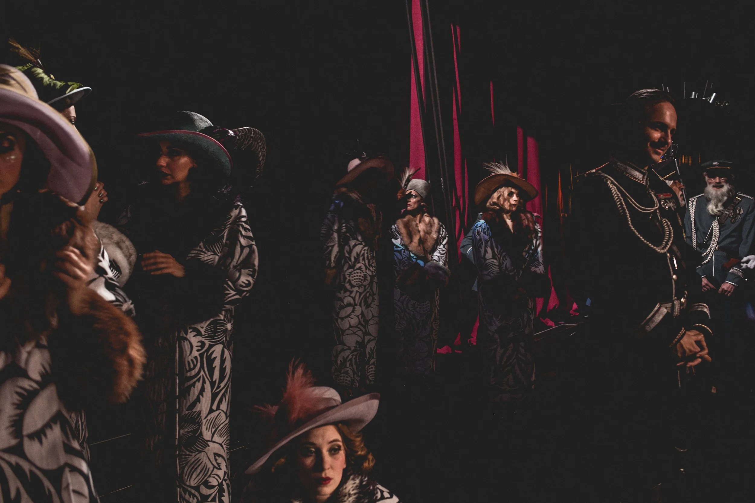 Damen und Männer in schwerer, auffälliger Kleidung und Hüten, wahrscheinlich in einer Theater- oder Modenschau, in einem dunklen Raum mit rotem Licht und schwarzem Hintergrund.