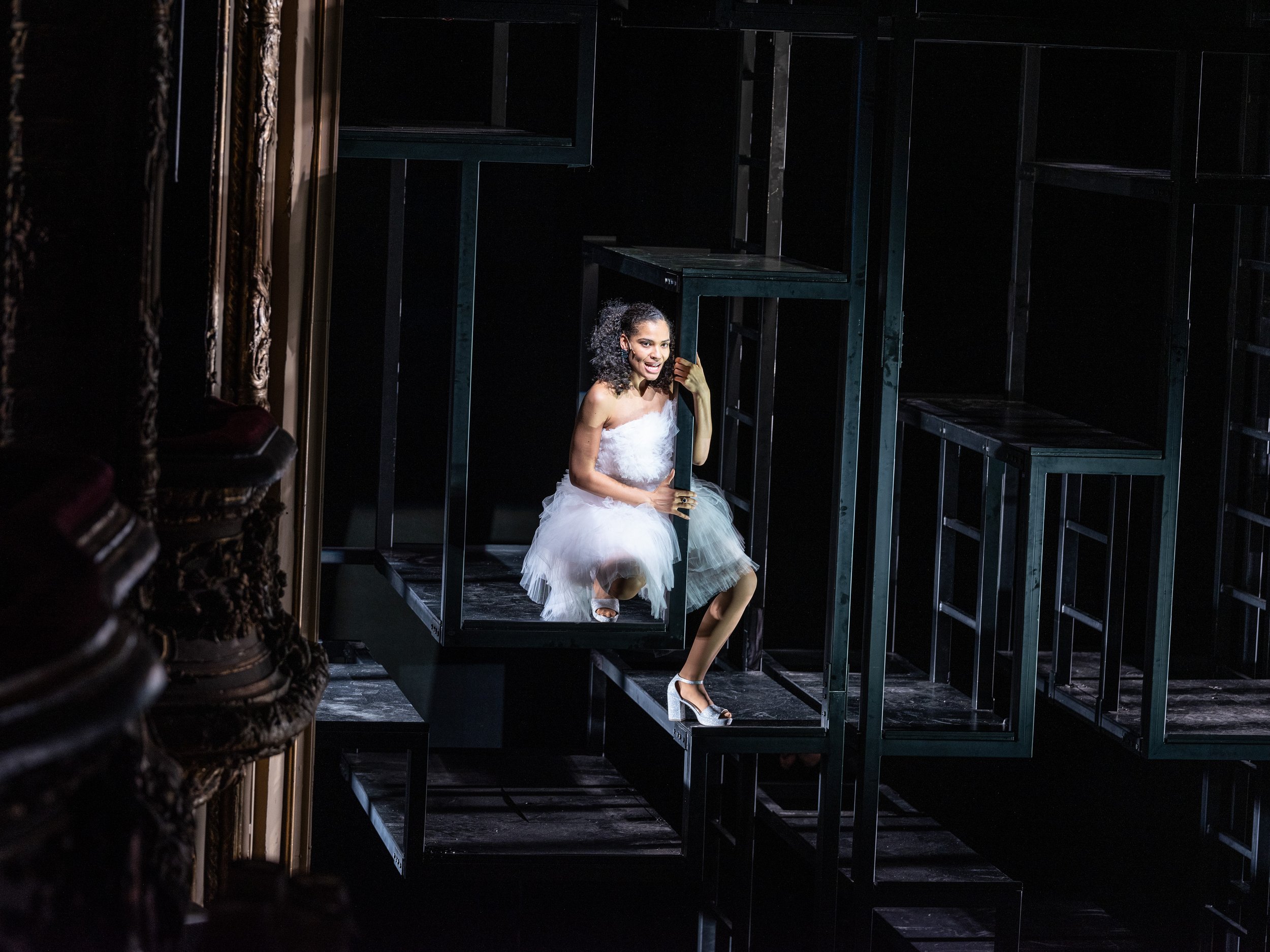 Eine Frau in einem weißen Tutu sitzt auf einem schwarzen Regalrahmen auf einer Theaterbühne, umgeben von weiteren schwarzen Regalrahmen. Sie lächelt und schaut nach oben.
