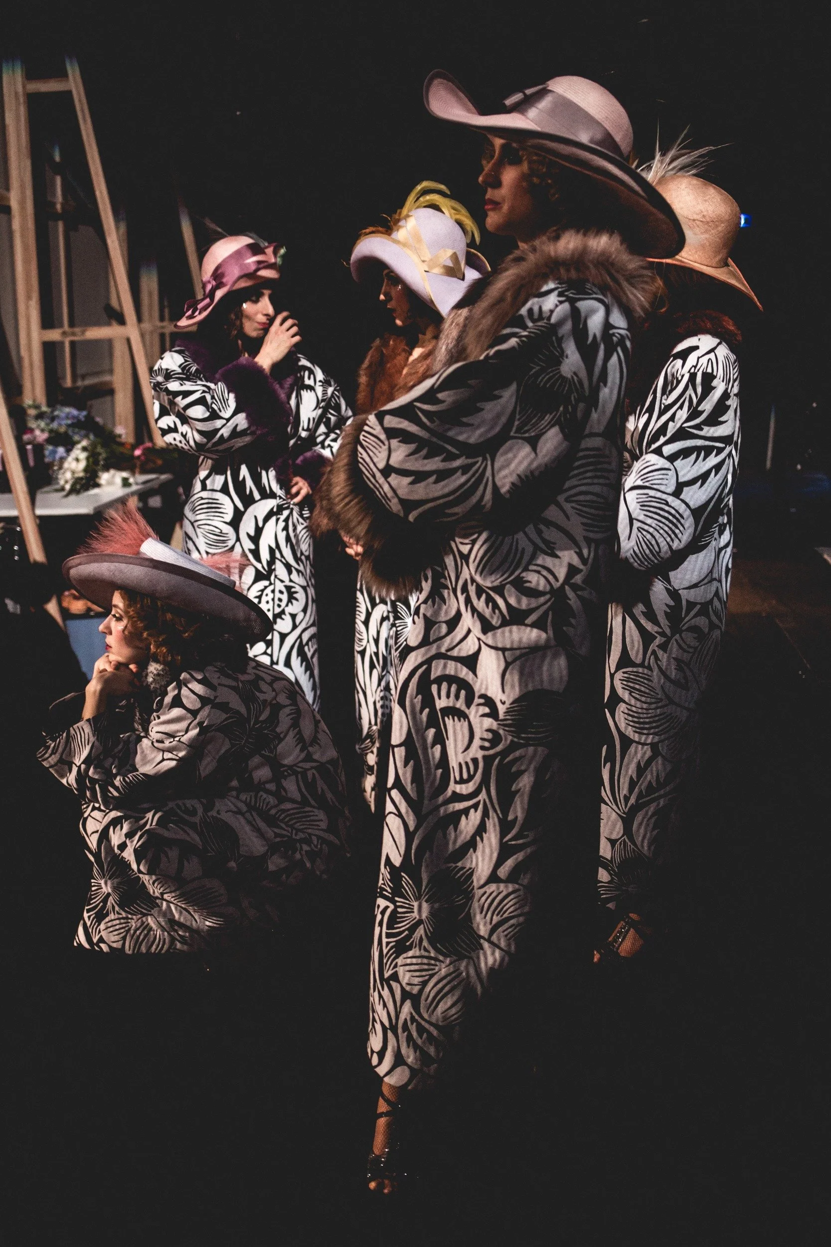 Fünf Frauen in modischer Kleidung und Hüten bei einer Modenschau oder Fotoshooting, im dunklen Raum.