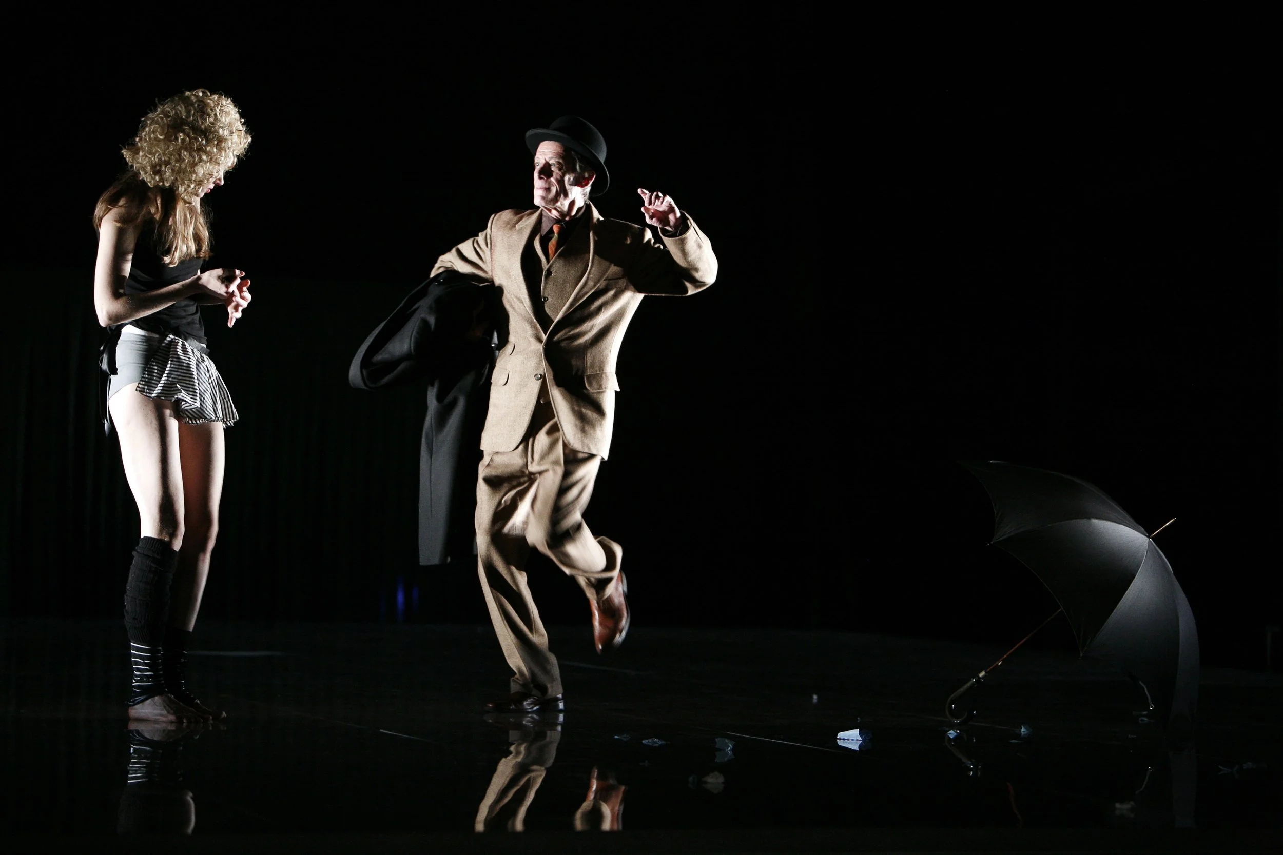 Zwei Schauspieler auf einer dunklen Bühne, einer trägt schwankend einen schwarzen Mantel und einen Hut, der andere ist in schwarzem Top, Shorts mit Streifen und hohen Socken, neben einem umgekippten schwarzen Regenschirm.