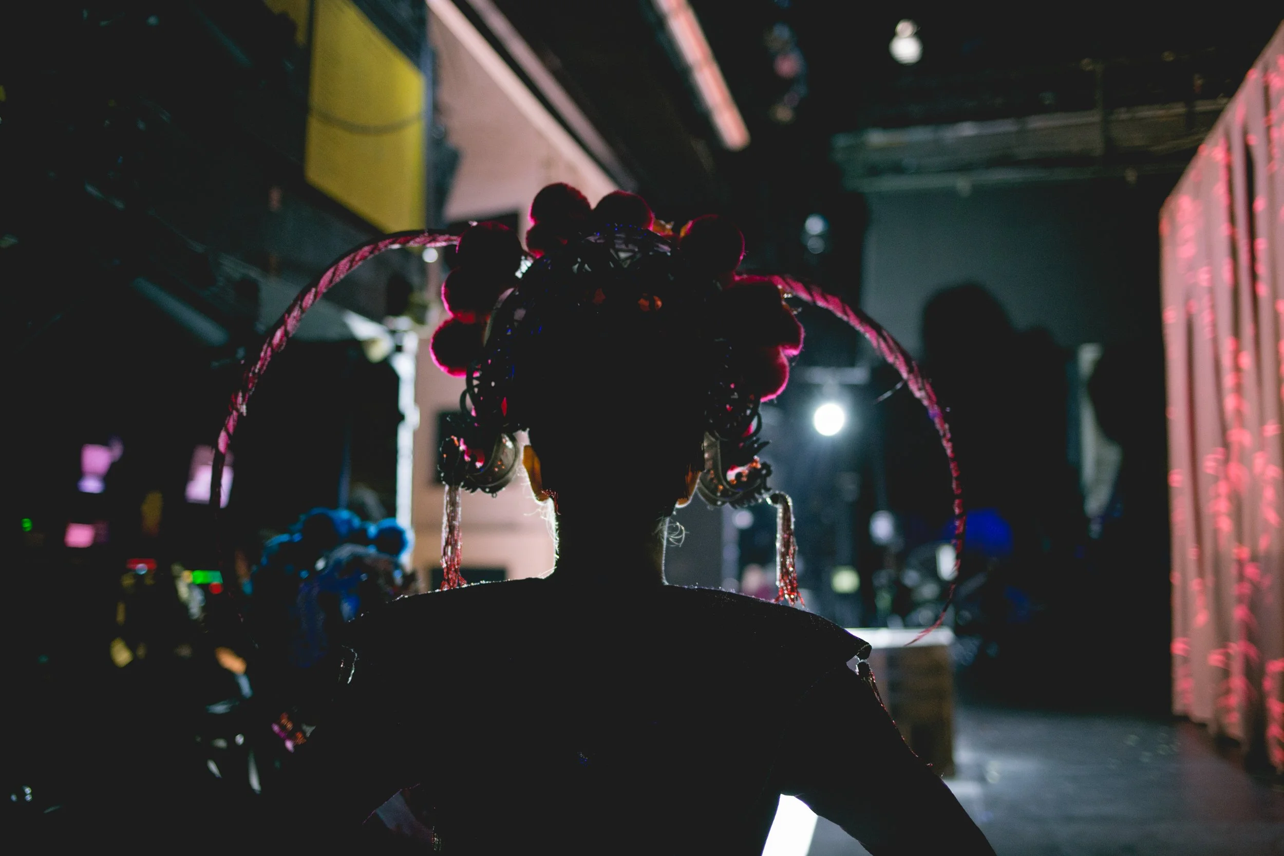 Eine Silhouette eines Frauenkopfes mit aufwendig geschmückter Frisur, beleuchtet von Bühnenlicht, im Hintergrund eine Bühne und zwei unscharfe Personen.