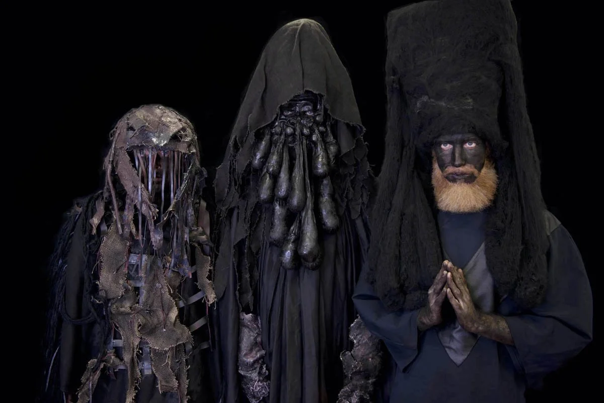 Drei Personen in dunkler, auffälliger Kostümierung, die an traditionelle, mystische Figuren erinnern, mit schwarz gefärbtem Gesicht und speziellen Masken vor dunklem Hintergrund.