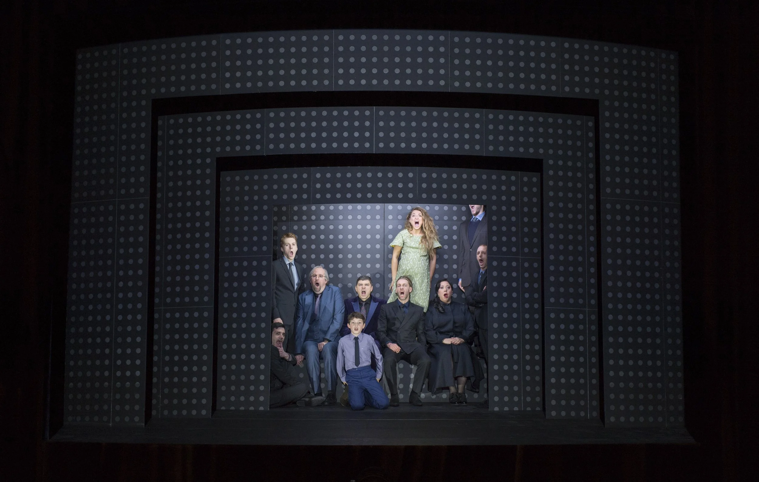 The image shows a group of actors on stage, with a woman in a green dress standing in the center, surrounded by others in formal clothing, all appearing to sing or perform together.