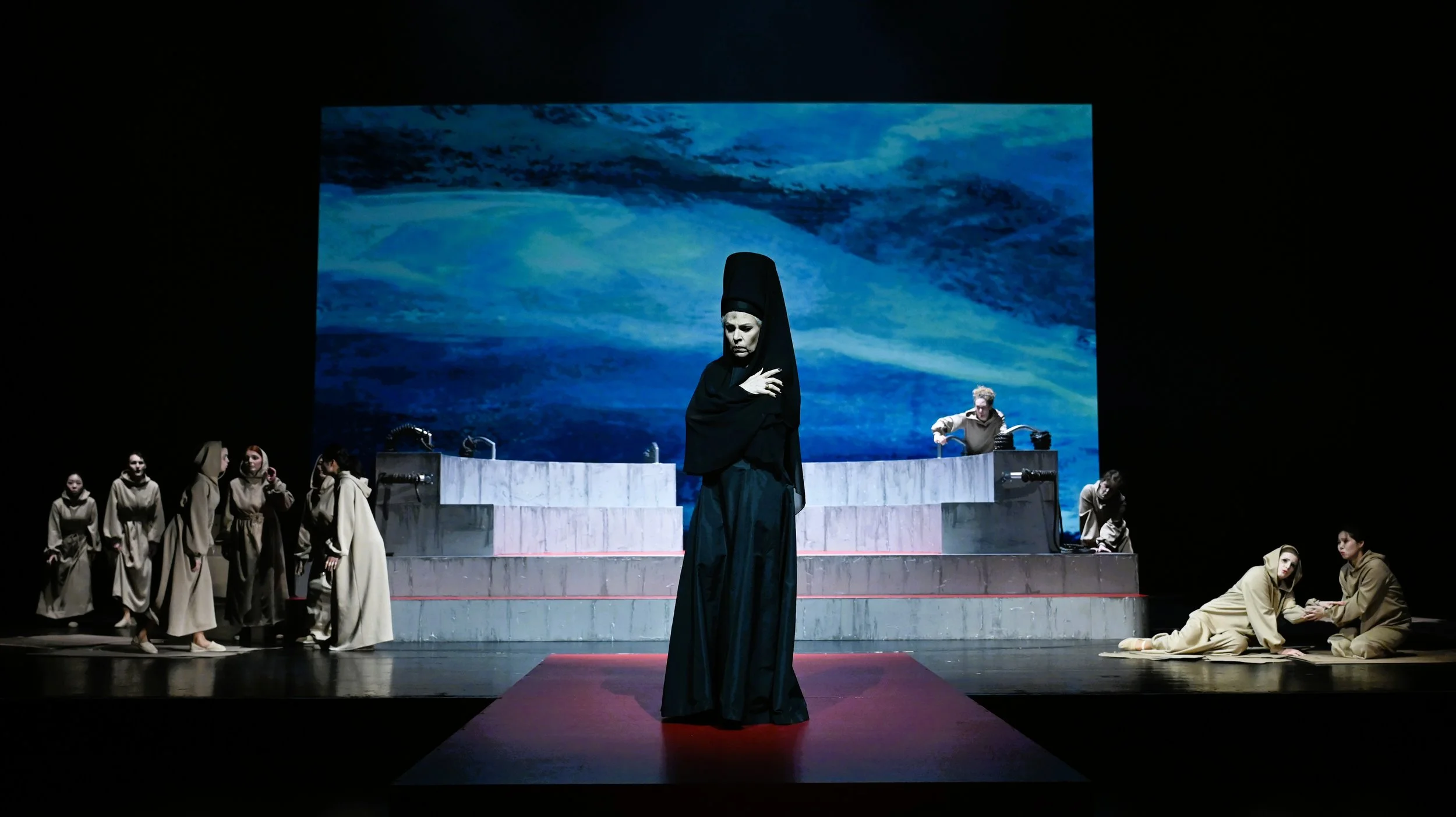 Bühnenaufnahme eines Theaterstücks mit mehreren Schauspielern in Kostümen, im Hintergrund eine große Leinwand mit einem stürmischen Meer. Im Vordergrund steht eine Person in dunklem Gewand mit einer hohen Kopfbedeckung.