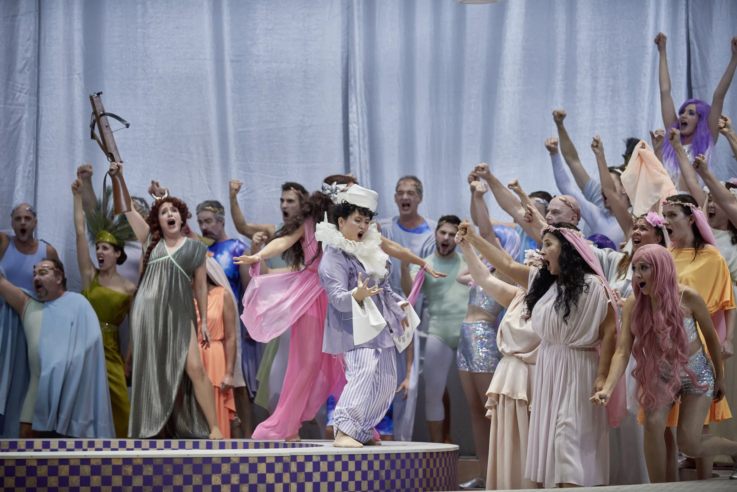 Szenenbild aus einer Theaterproduktion mit einer großen Gruppe von Schauspielern in bunten Kostümen, die laut und energisch auf der Bühne agieren, darunter eine Frau mit rosa Haaren und ein Mann in einem alten, viktorianischen Kostüm, die alle leiden